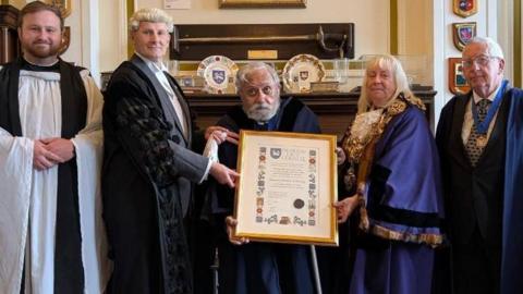 Official group shot of Jonathan Saksena in the centre, surrounded by the mayoress and another official - both in ceremonial blue robes - on the right. On the left is a clergyman in the traditional white surplice tunic and black scarf alongside a man wearing a judge's white wig plus black robe. Councillor Saksena is wearing a dark ceremonial robe and holding a walking stick as he also holds a frame showing his Freedom of Preston certificate.