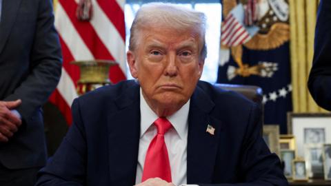 Donald Trump sitting at his desk wearing a dark suit and red tie.