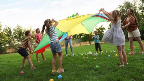 A group of young children dressed in summer clothing, gathered in a circle on the grass, surrounded by a line of trees. Plastic balls in bright colours lie on the grass by their feet. The children are raising a sheet which is divided into colourful sections, above their heads as part of a game. They are joined by a female adult. 