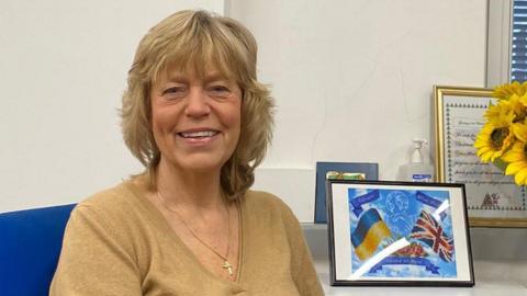 A woman with blonde hair. She is sat in a blue chair in a white room, smiling at the camera. She is wearing a light brown top. To her left, our right, is a framed picture showing the UK and Ukrainian flags.