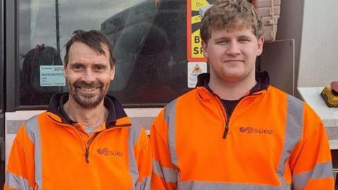 Two men in orange hi-viz suits stand in front of a bin lorry.