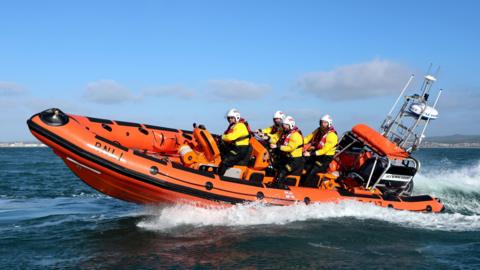 An orange RNLI boat with five people on board, all wearing white helmets and yellow jackets, is out at sea. 