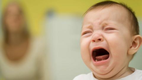 Baby crying with a mother out of focus in the background.