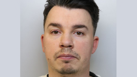A police mugshot of a man in his early 30s with short dark hair, dark stubble and brown eyes. He is wearing a black T-shirt underneath a white jumper, and standing against a grey background. 