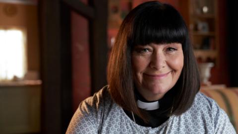 Dawn French in character as the Vicar Of Dibley, - The Reverend Geraldine Granger. She has a black bob haircut, dog collar and is wearing a white top. She is smiling.