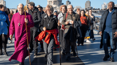 A group of people in religious clothing wearing crosses and carrying sticks crossing bridge with over Thames with St Paul's Cathedral in background 