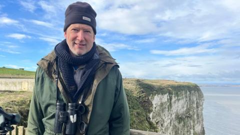 A man wearing warm closing stands on a platform in front of tall chalk cliffs overlooking a grey-blue sea under a cloudy blue sky. He is wearing a black woolen heat, a navy-blue scarf, a free coat and binoculars.