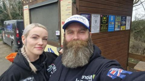 Amber wearing a black hoodie and Adam wearing a black hoodie- smiling for a selfie in front of their burger shed.
