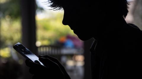 A teenage boy in silhouette is holding a phone. He is outside.