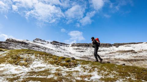 Man hiking in hills with patchy snow laden and blue, sunny skies overhead