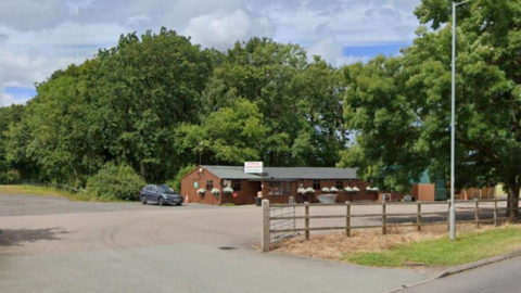 A long brown building, set back from the road, with a grey roof. There are large trees behind the building and a large concreted area for cars.