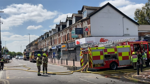 Three firefighters in protective clothing and some wearing helmets are on the right next to the cab of a fire engine. Two others are talking in the street in the centre of the photo, with yellow hoses trailing from the back of the fire engine across what looks a typical high street