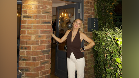 A woman with blonde hair, wearing a brown halter-neck top and white trousers, leans in a doorway and smiles at the camera.
