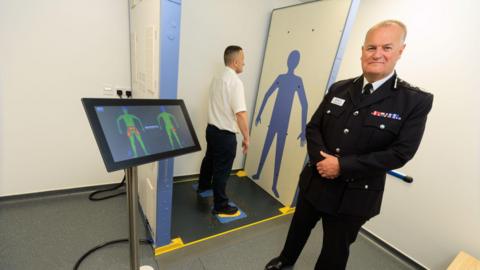 A man can be seen standing in the bay of a security scanner with his feet on the marked spots and his arms held out to the sides of his body. In the foreground on the right is Chief Constable Sir Stephen Watson looking at the camera. On the left is a monitor showing the results of the body scan.