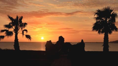 The sun rises over a ruined building in front of the sea. The sky is glowing orange over head. On either side of the building are two palm trees.