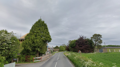 A streetview image of the the A4361 in Winterbourne Monkton. The two-lane road runs past a small village. There is a green field on the right and trees and hedgerows on both sides of the pictures, with some houses and farm buildings. The sky is overcast.  