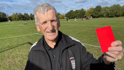 87-year-old football referee Percy Parker stands on a grassy pitch, holding up a red card towards the camera. He is wearing a black Adidas jacket with an FA referee badge.
