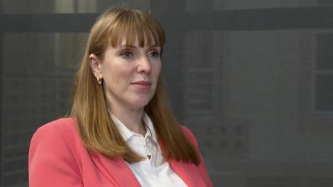 Deputy Prime Minister Angela Rayner sitting in a corridor at the BBC's studios in Millbank, Westminster. She wears a white blouse and a pink blazer.