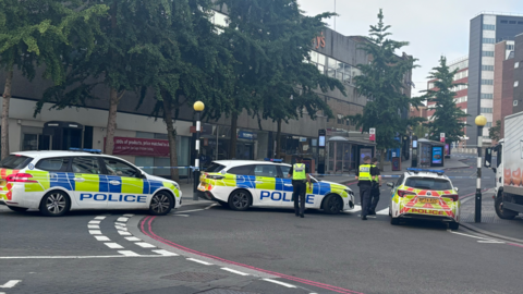 A cordoned off street with police cars and  tape sectioning off a road. There are closed shops on either side of the road in the background. There are also some trees on the left side pavement in the distance. 