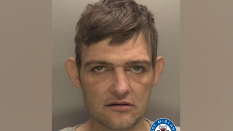 Topham has short dark hair in his police mugshot and is wearing a light T-shirt.