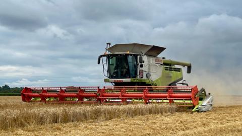 A green combine harvester at Waltham St Lawrence is cutting a field of golden crops. There is a large red cutting blade across the centre of the picture. Above the sky has grey clouds.