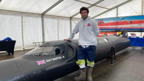 Guy Martin is wearing a grey hooded sweatshirt, blue trousers and dark wellies. He is standing by a long grey vehicle that will be used to attempt to break the 400mph barrier.