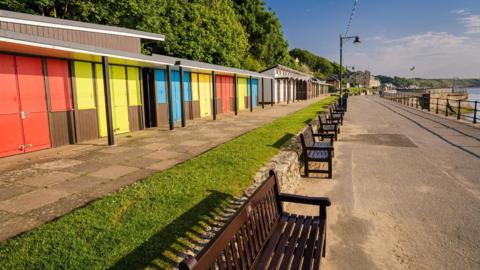 A row of brightly coloured beach huts. In front of the huts are several wooden benches which are overlooking the sea. 