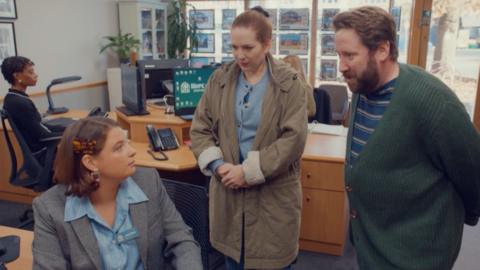 Characters Rachel and Paul Jessop, looking concerned, stand talking to an estate agent in her office