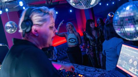 A woman with short blonde hair is seen DJing in a dark room, while people dance behind her