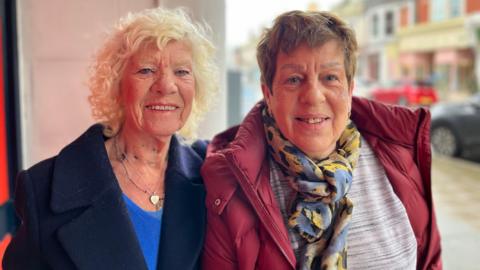 Valerie Cartwright, left, and Shirley Percival standing outside the Buzz Bingo in Worthing. Valerie is wearing a navy blue woolen coat and Shirley is wearing a red jacket and floral scarf. Both are smiling at the camera