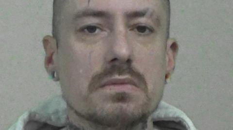 Mugshot of Phillips. He has a short beard and shaved head, with tattoos on his neck and large round earrings in the lobes of his ears