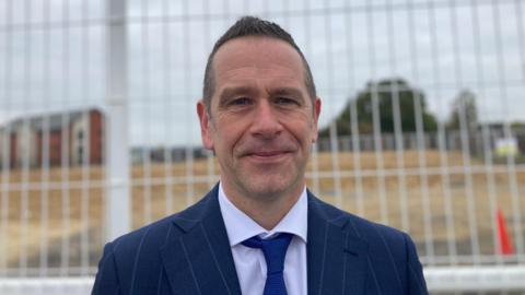 A man in a suit looking at the camera with a soft smile. He is wearing a navy striped suit and standing in front of a building site with a gate behind him. 