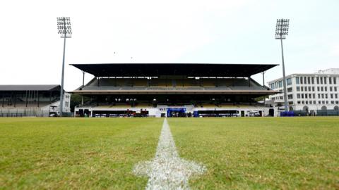 A view of a two-tiered sport stadium stand. In the foreground is a green sports pitch marked with white paint to indicate its centre spot.
