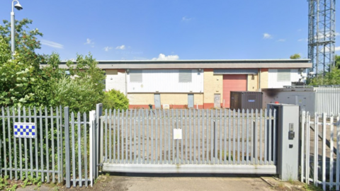Industrial unit on a business park in York with blue sky above, bushes and CCTV to the left, and a metal fence and gates in front of the red and yellow brick building.