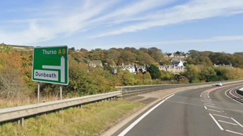 A green road sign on the A9 with arrows pointing north to Thurso and west to Dunbeath. A few metres along the road from the sign are houses of Dunbeath.