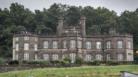 A large grey building with a crenellated roof line, a turreted central tower and white sash windows.