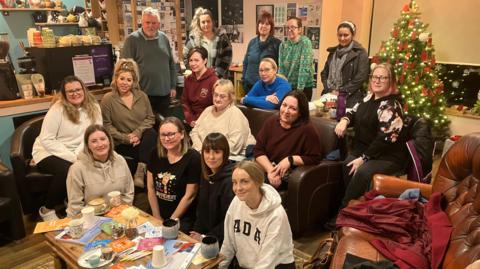 A group picture of sixteen people, mainly women, with a Christmas tree on the right hand side.