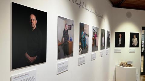 A number of rectangular portraits of staff and volunteers of Les Bourgs Hospice displayed on a plain white wall. They each have an informational plaque below each one.