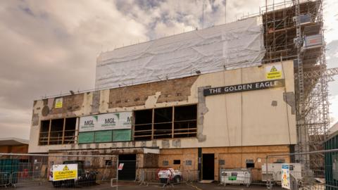 The exterior of the former Golden Eagle hotel. Cream plaster and paint on the front is crumbling, while chunks have been taken out near the rear. Scaffolding has been erected at the front and over the top of the building.