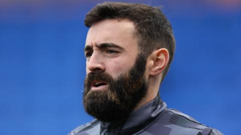 Jack Price, wearing a tracksuit and with a dark beard, against a blue background, looks forward