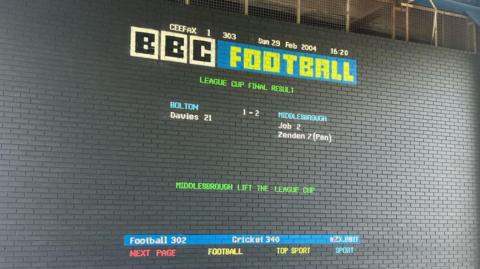 Mural in the style of the former television teletext service Ceefax. Set against a dark grey brick wall it states in red, yellow, green, blue and white letters: BBC Football: League Cup Final Result. Bolton 1 -2 Middlesbrough. Middlesbrough lift the league cup