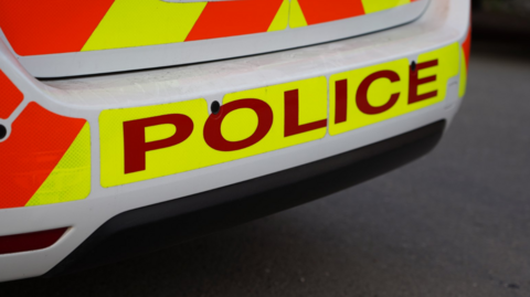 Rear bumper of a police car, which reads 'police'. The car has reflective yellow and orange stickers.