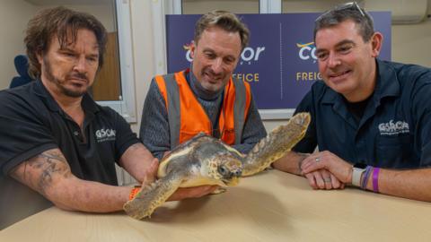 Three men sat around a table with the man on the left holding a loggerhead turtle. The turtle, called Barnacle Bill is waving bye with its right fin. 