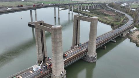 An aerial view of the Kingsferry bridge, seen in the foreground of the picture and which has large concrete pillars in two places which lift the middle section of the bridge which runs over a river. In the background and higher up the road bridge is also seen.