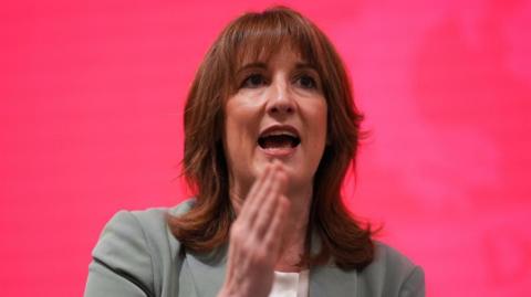 Rachel Reeves speaking in front of a red backdrop. She has shoulder length brown hair, her right hand is raised and she's wearing a light green coloured suit.