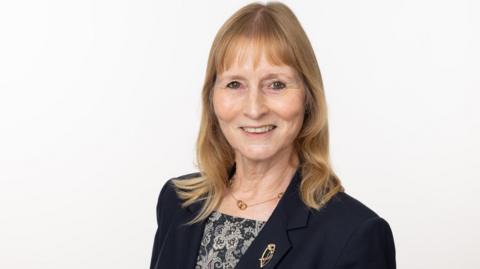 Barbara Corbett is smiling and has shoulder length blonde hair and is wearing a navy jacket. 