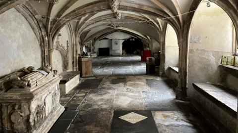 The inside of the crypt. Vaulted cielings can be seen and there are tombs on the left side of the stone crypt. Different coloured flagstones can be seen on the floor.