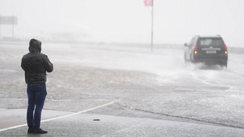 A car driving through floodwater in Galway which a person looks on. 