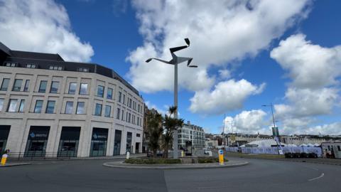 A tall silver column, with the three legs of mann, stands above a roundabout. There is a modern light building in the background and marquis tents on the other side.