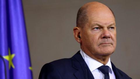 German chancellor Olaf Scholz wearing an earpiece and standing in front of the EU flag